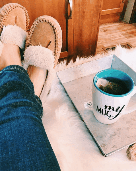 Mug of tea on tray next to woman wearing moccasin style slippers sitting inside RV.