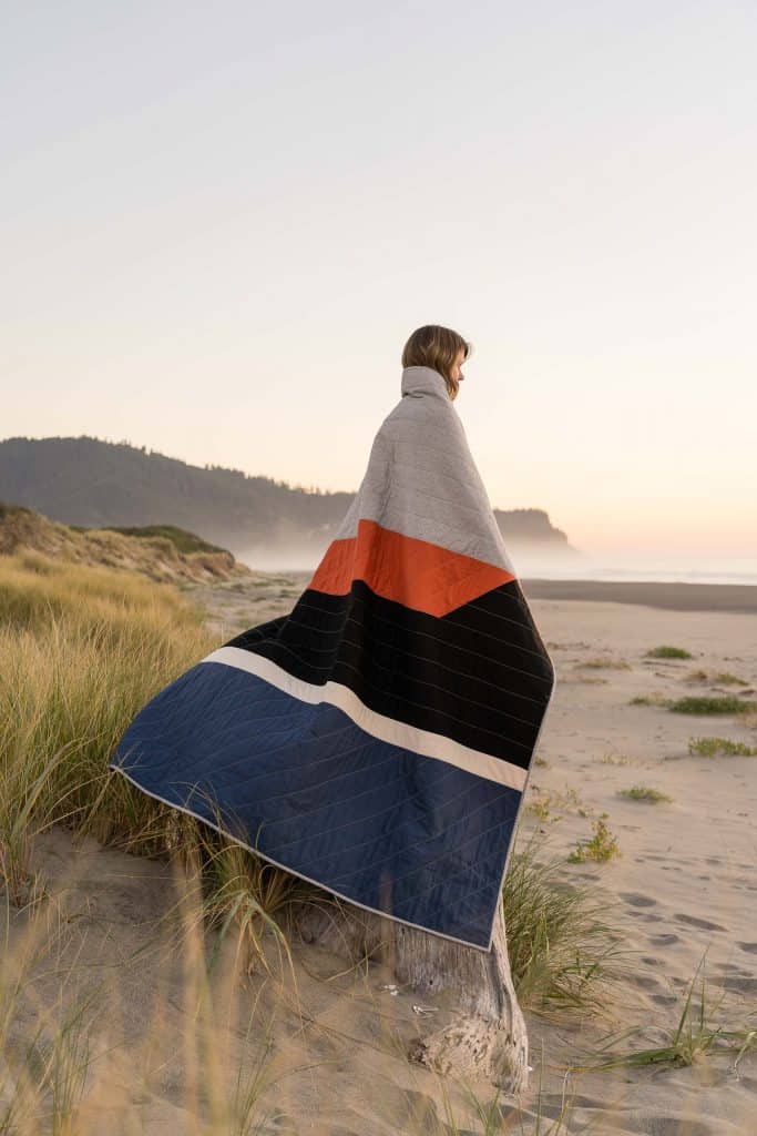Woman on beach holding large quilted blanket she learned to craft and sew while RVing