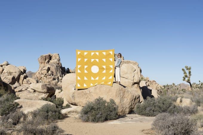 Laura Preston displaying  a large quilt while standing on a rock formation at an RV stop