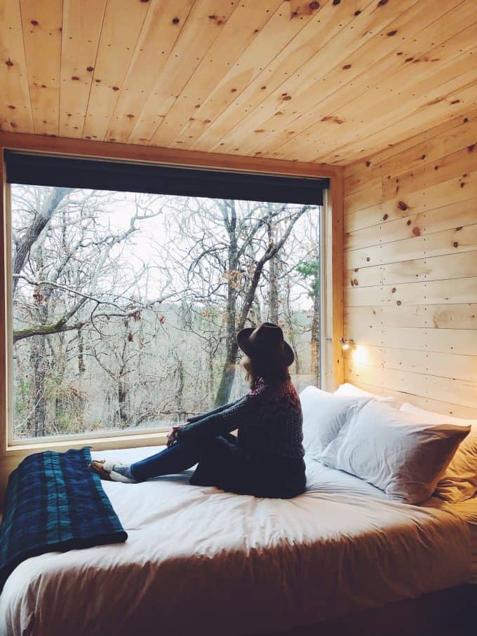 Woman looking through window of tiny home toward outside wooded area