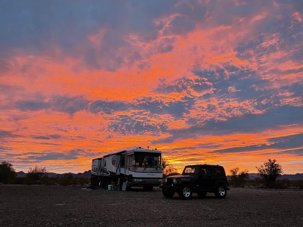 Jeep parked in front of large RV with bright orange sunset.