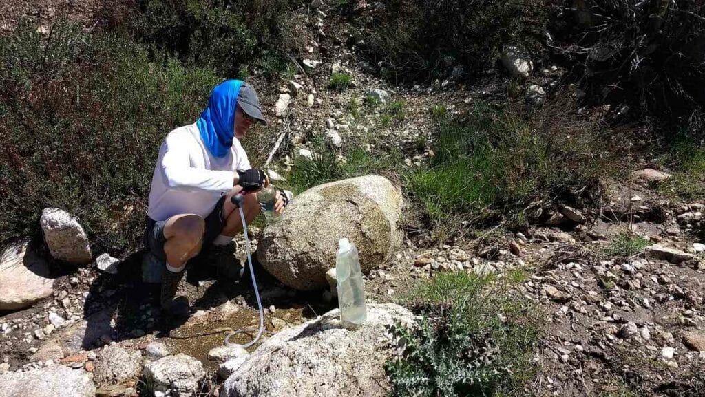 Male hiker kneels next to stream to filter water while on hike.