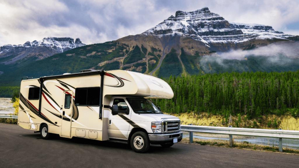 Cream colored motorhome RV parked near guardrail with snow capped mountains in background.