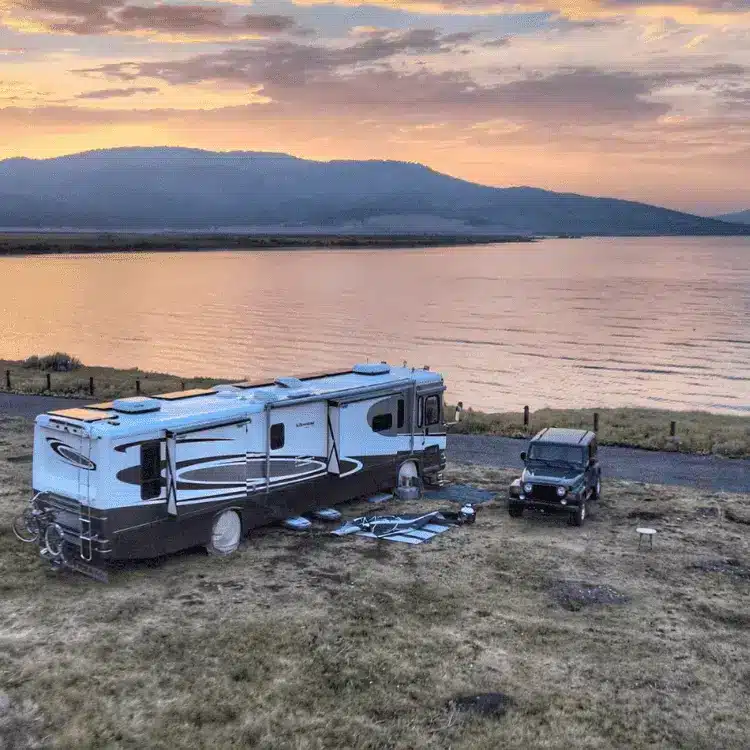 Class A RV and Jeep parked in boondocking area near lake with sun setting behind distant mountains.