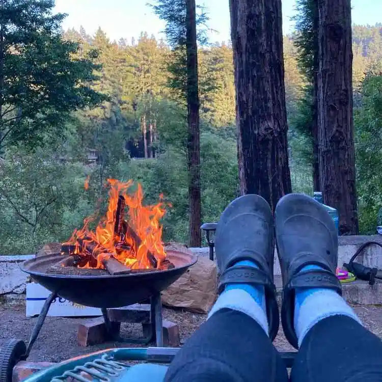 Wood burning campfire contained within a portable, elevated fire pit with photographer's feet in foreground and forest trees in background