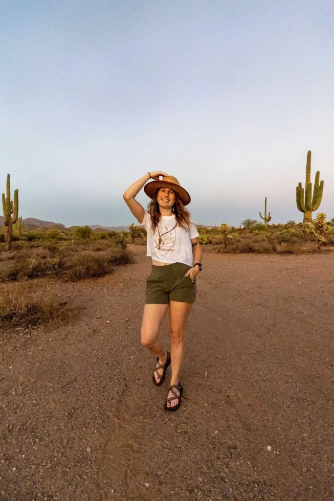 Woman holding onto top of hat, wearing hiking shorts and walking toward camera