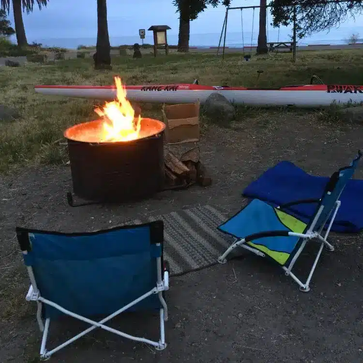 Camp chairs set up next to wood burning campfire that is contained within a metal drum near playground equipment and shores of the beach