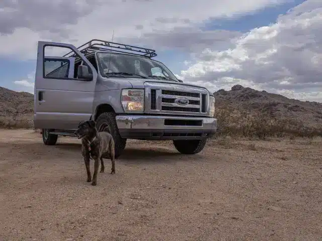 Brown dog walking in front of campervan with passenger door open parked in open desert area.